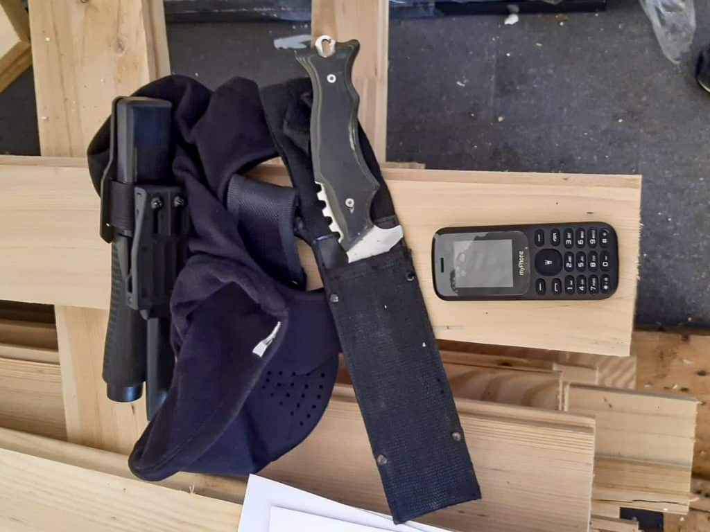 Na zdjęciu widoczny pistolet, maska na twarz, pasek do broni oraz telefon komórkowy na drewnianym blacie.