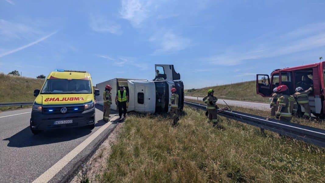 Wypadek ciężarówki na drodze S7, pojazd leży na boku na poboczu, wokół strażacy i karetka.