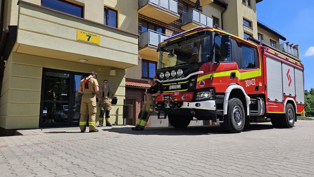 Straż pożarna i ratownicy przed wejściem do budynku na osiedlu w Olsztynie, obok czerwony wóz strażacki.