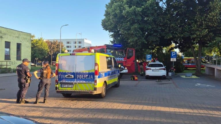 Na parkingu przed komendą policji stoją radiowozy, straż pożarna i samochód osobowy, w tle drzewa i budynki.