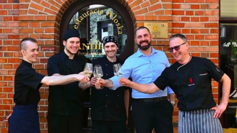 Grupa kucharzy i właścicieli restauracji stojąca przed wejściem do lokalu, trzymająca kieliszki z winem.