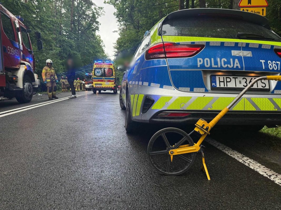 Na drodze krajowej nr 16 w Zawadach Małych stoi radiowóz policji, w tle służby ratunkowe i uszkodzone samochody po zderzeniu.