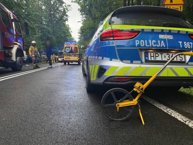 Na drodze krajowej nr 16 w Zawadach Małych stoi radiowóz policji, w tle służby ratunkowe i uszkodzone samochody po zderzeniu.
