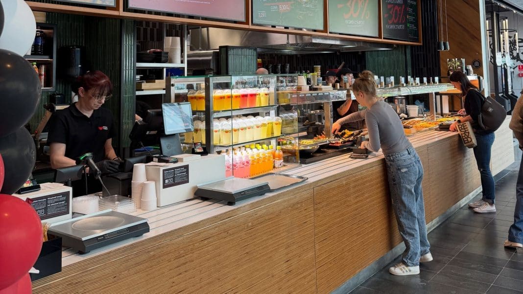 Kolejka klientów przy bufecie z napojami i jedzeniem w strefie food court galerii handlowej.