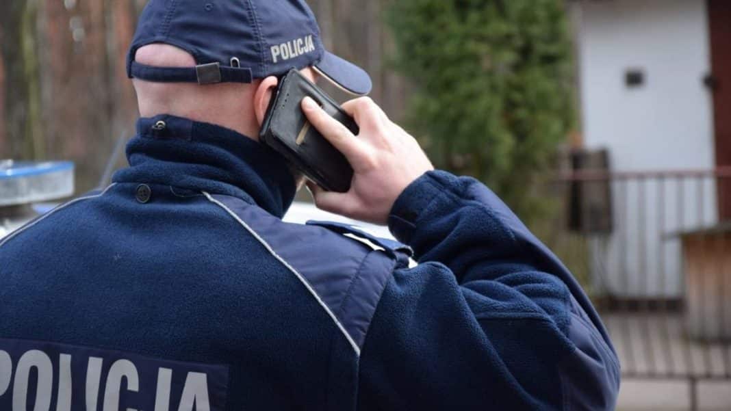 Policjant rozmawia przez telefon na zewnątrz, w tle drzewa i budynki.