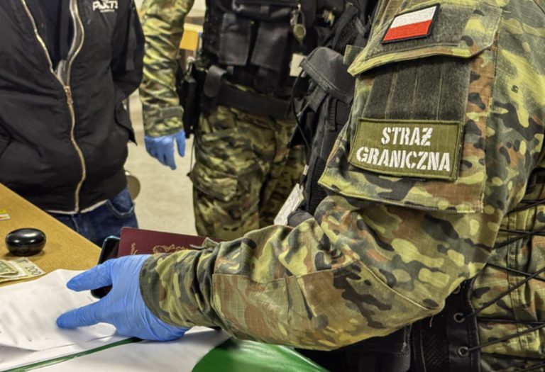 Funkcjonariusz Straży Granicznej w kamuflażu z napisem 'Straż Graniczna' podczas kontroli, obok osoby w ciemnej kurtce.