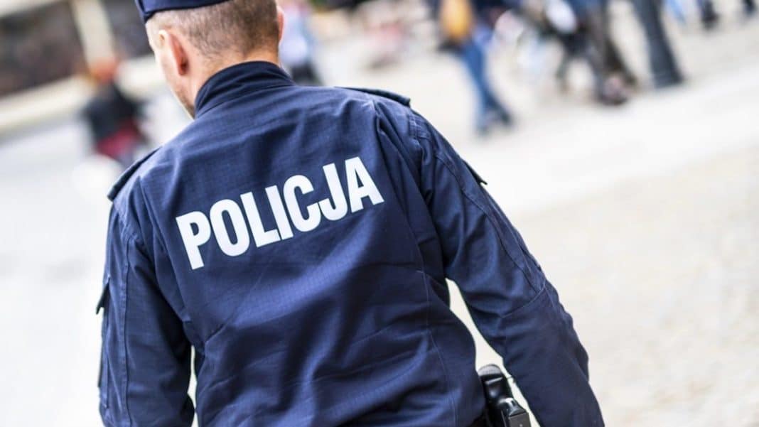Policjant w niebieskiej mundurze z napisem 'POLICJA' na plecach, widziany od tyłu na ulicy.