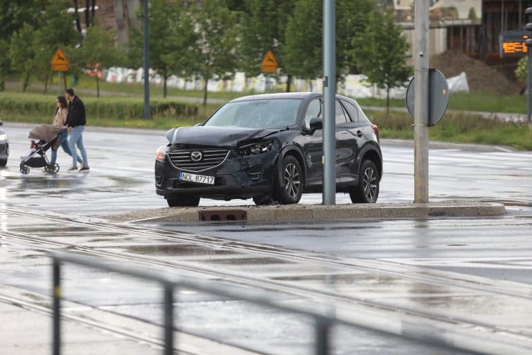 Uszkodzony czarny SUV na skrzyżowaniu, zderzenie z innym pojazdem, mokra nawierzchnia, w tle piesi i trawa.