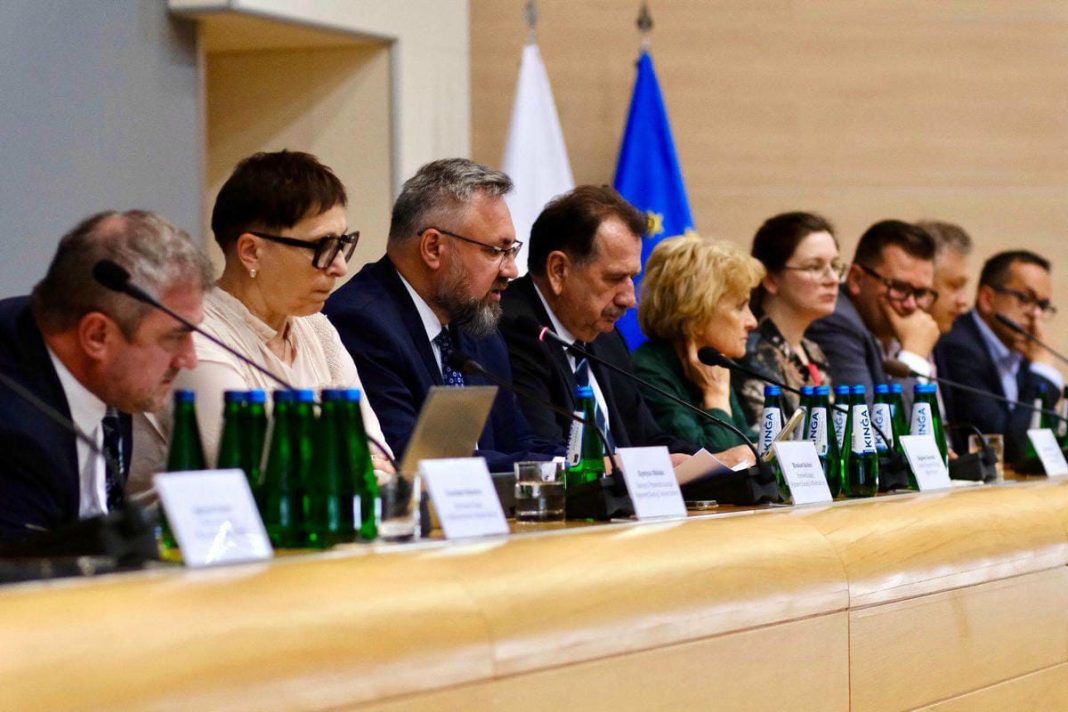 Grupa osób siedzi przy stole podczas konferencji prasowej, z flagami w tle.