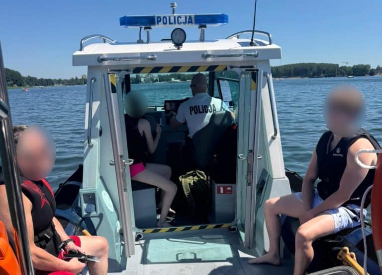 Policja na łodzi patrolowej na jeziorze Ukiel, w tle woda i linia brzegowa z drzewami.