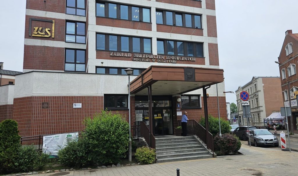 Budynek olsztyńskiego ZUS z widocznym napisem i wejściem, na zewnątrz kilka osób, pochmurny dzień.