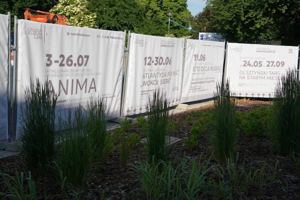 Płot przy Wysokiej Bramie w Olsztynie z panelami informacyjnymi i roślinami w tle.