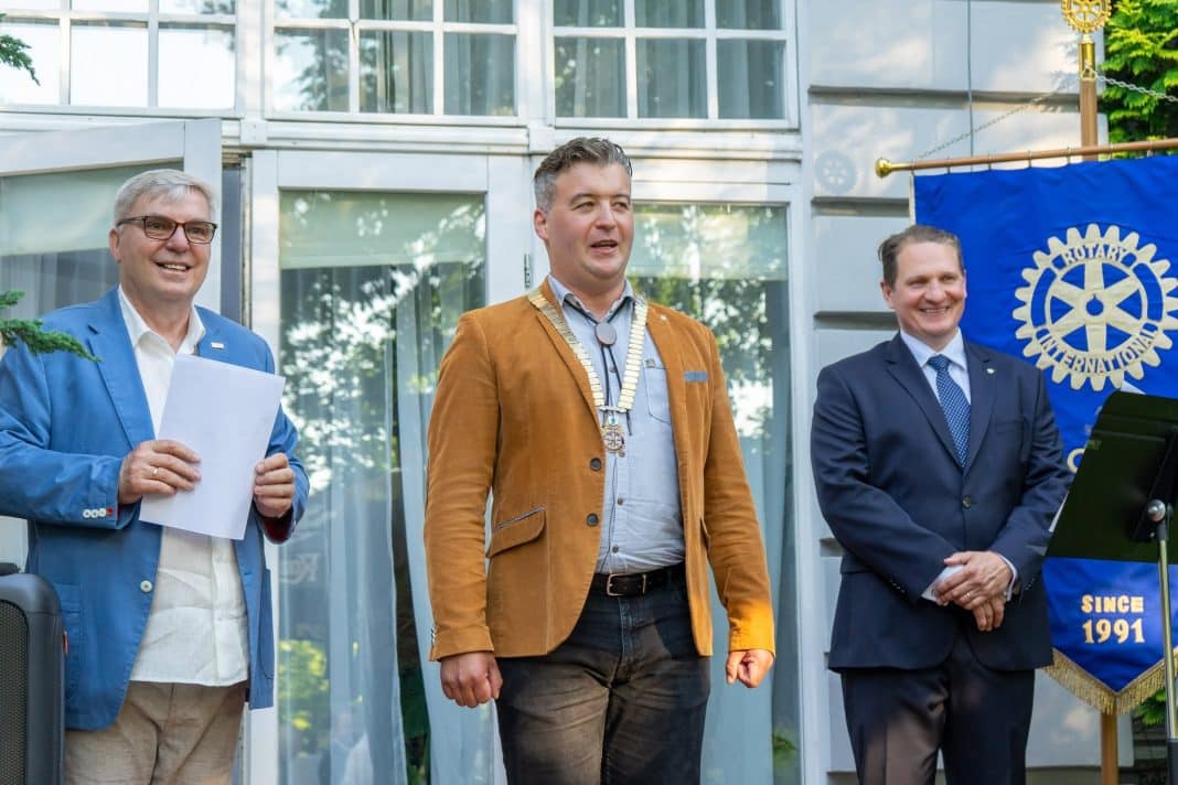 Trzech mężczyzn na uroczystości Rotary Club Olsztyn, jeden z nich z medalem, w tle flaga organizacji.