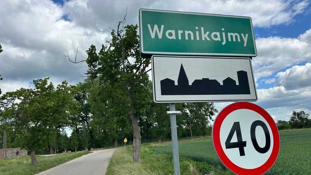 Zdjęcie znaku wsi Warnikajmy na tle drzew i nieba z chmurami.
