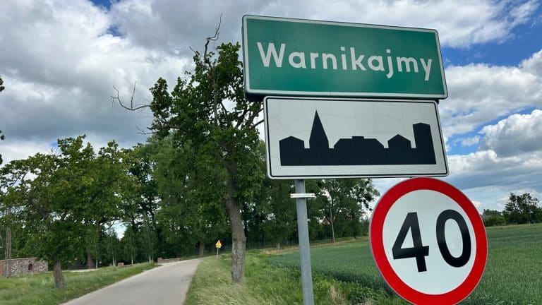 Zdjęcie znaku wsi Warnikajmy na tle drzew i nieba z chmurami.