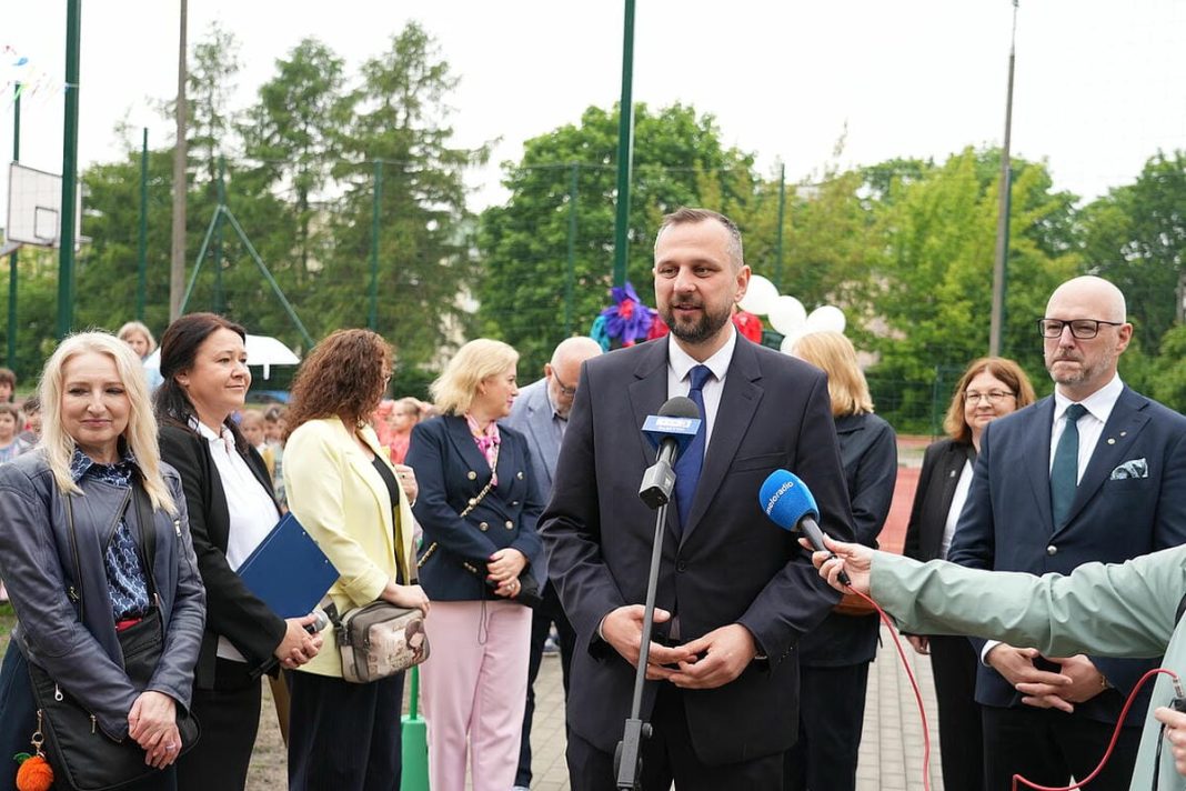 Grupa osób na boisku szkolnym podczas otwarcia nowoczesnego kompleksu sportowego w Olsztynie.
