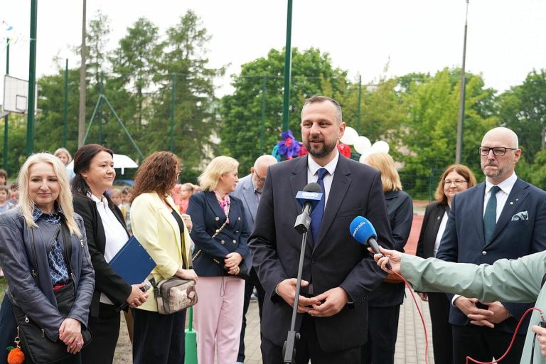 Grupa osób na boisku szkolnym podczas otwarcia nowoczesnego kompleksu sportowego w Olsztynie.