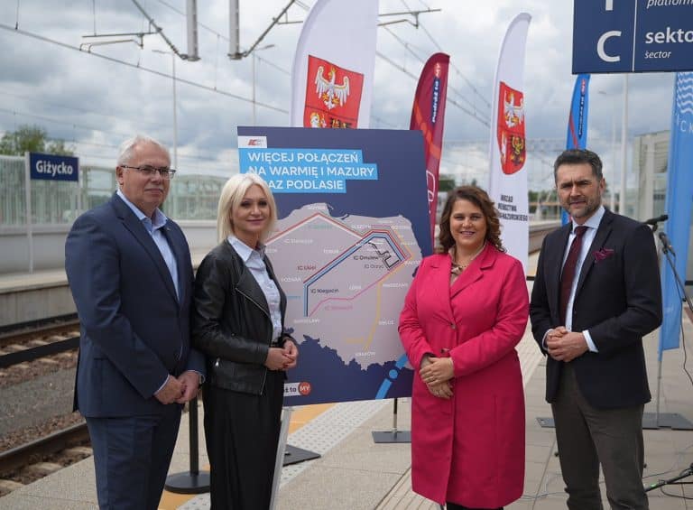 Grupa czterech osób stojących na peronie kolejowym obok planszy z mapą połączeń kolejowych w regionie.