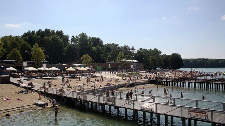 Plaża nad jeziorem Ukiel z ludźmi opalającymi się i kąpiącymi się, w tle zieleń drzew i pomosty.
