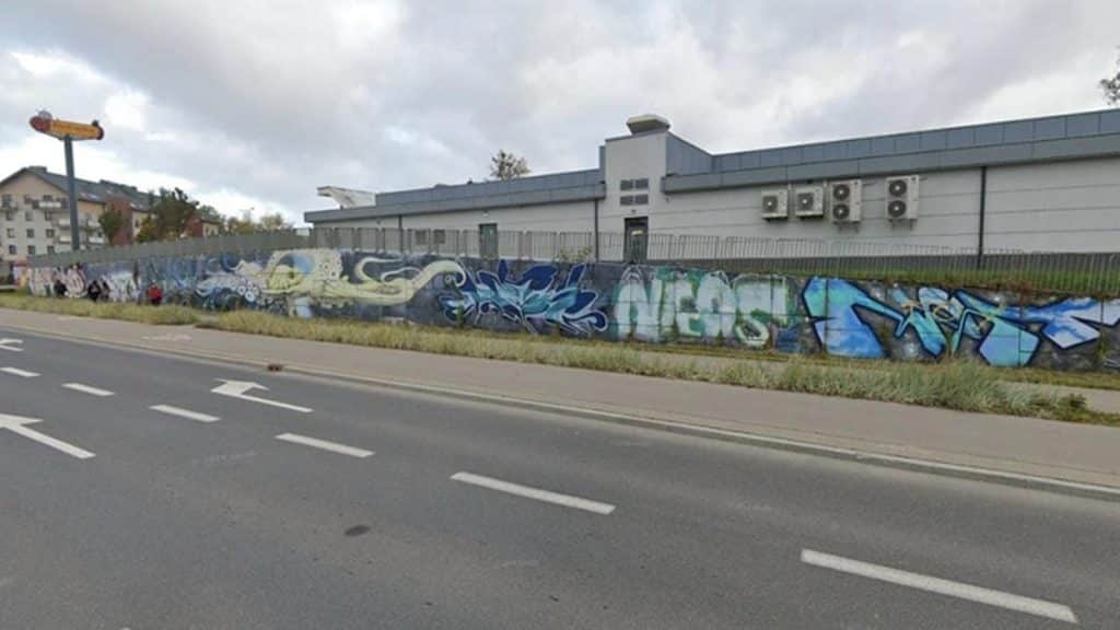 Murale graffiti na betonowej ścianie przy ulicy w Olsztynie, w tle budynek i chmury na niebie.