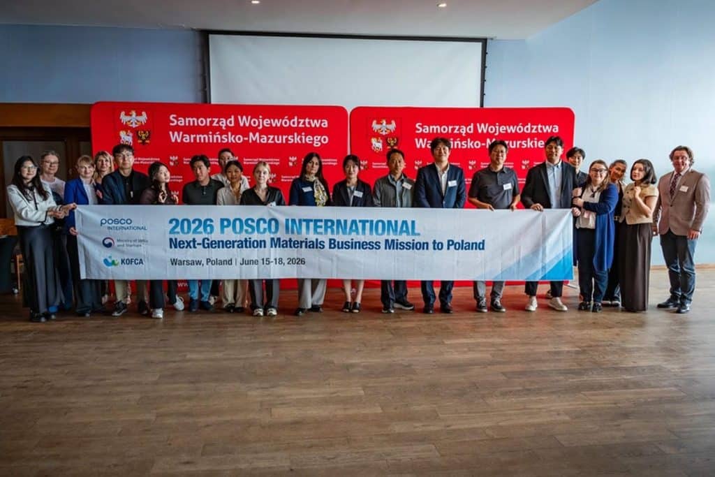 Grupa osób na spotkaniu biznesowym z banerem dotyczącym misji gospodarczej w Warszawie, w tle czerwone tablice z napisem "Samorząd Województwa Warmińsko-Mazurskiego".
