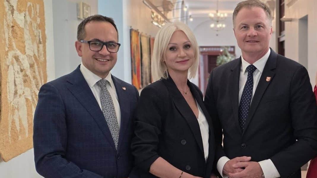 Trzy osoby w formalnych ubraniach stoją w eleganckim wnętrzu, na tle obrazów i ozdobnych elementów.