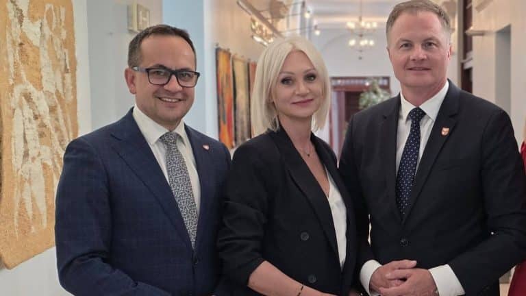 Trzy osoby w formalnych ubraniach stoją w eleganckim wnętrzu, na tle obrazów i ozdobnych elementów.