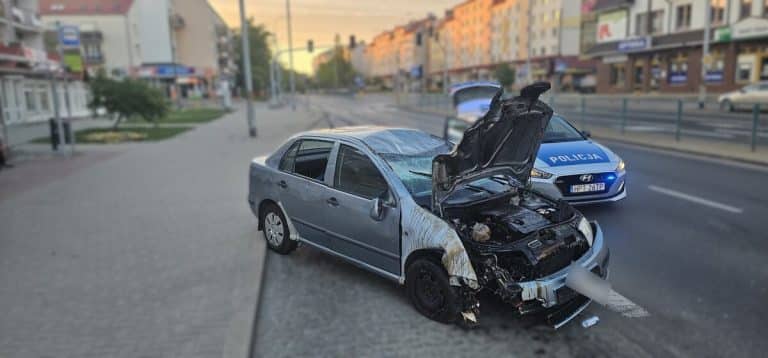Uszkodzony samochód osobowy po wypadku na ulicy, przewrócony na bok, z widocznymi uszkodzeniami silnika i karoserii.