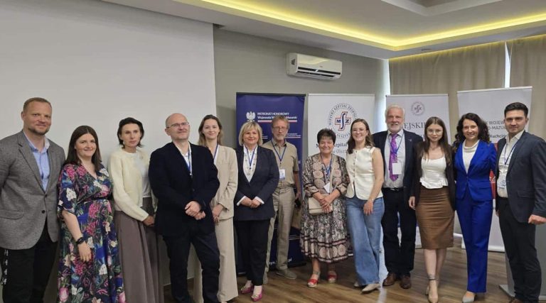 Grupa osób na konferencji w sali, stoją przed planszami i banerami.