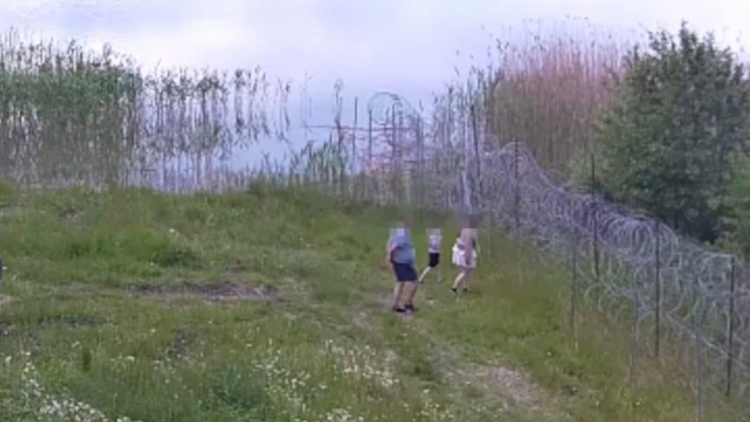 Grupa turystów w strefie nadgranicznej przy jeziorze Granicznym, blisko ogrodzenia i tablic ostrzegawczych.