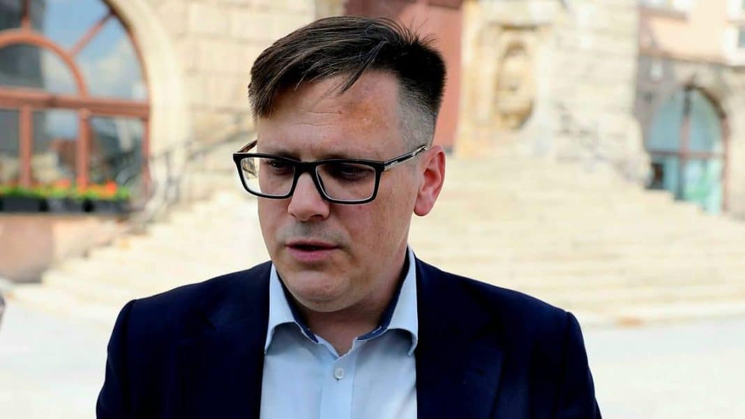 Mężczyzna w okularach i garniturze na tle budynku ratusza, podczas protestu przed urzędem.