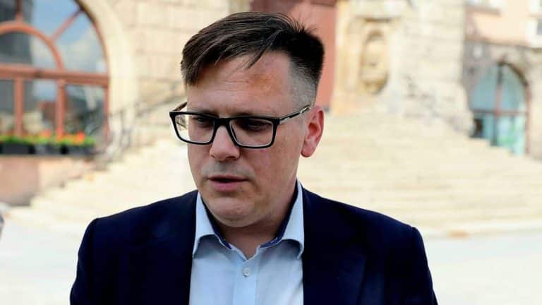 Mężczyzna w okularach i garniturze na tle budynku ratusza, podczas protestu przed urzędem.