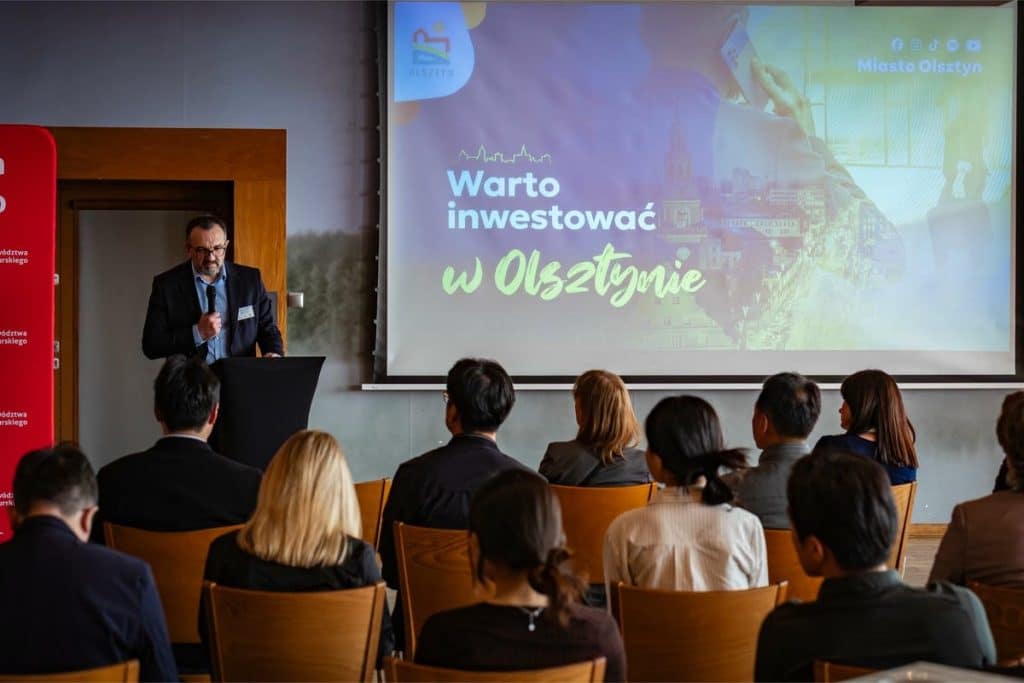 Prezentacja podczas spotkania biznesowego w Olsztynie, na ekranie napis 'Warto inwestować w Olsztynie' oraz grupa słuchaczy.