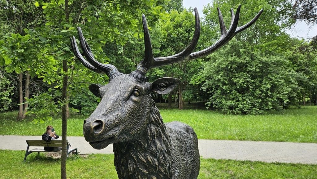 Rzeźba jelenia szlachetnego w parku, ludzie na ławce robią zdjęcia, otoczenie zielenią.