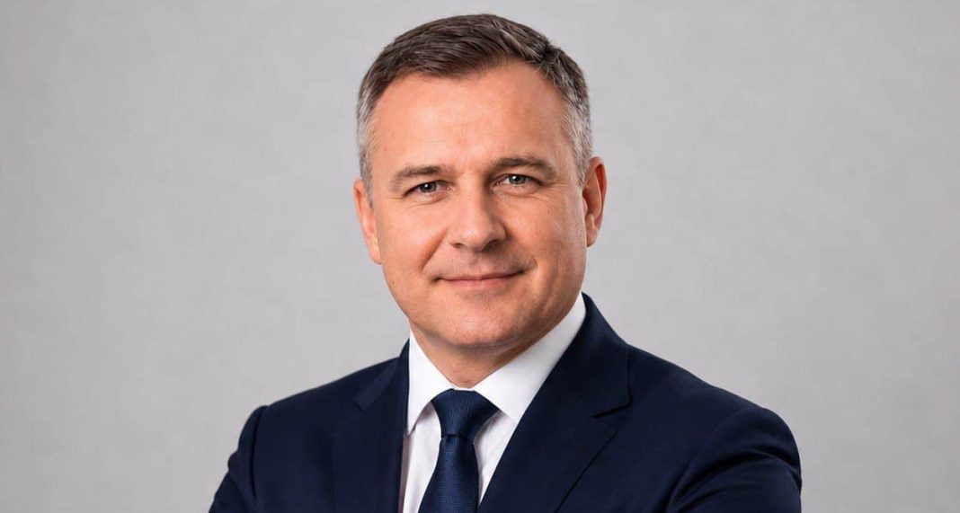 Mężczyzna w ciemnym garniturze, uśmiechnięty, na neutralnym tle.