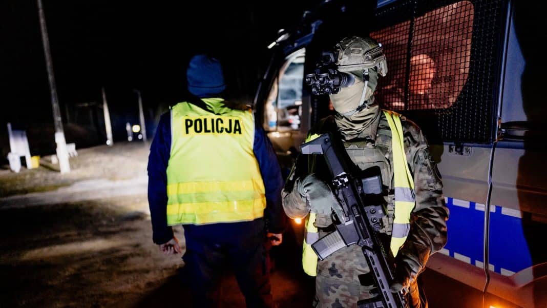 Żołnierz z bronią i kamizelką w kamuflażu, obok policjant na nocnym patrolu, w tle pojazd wojskowy.
