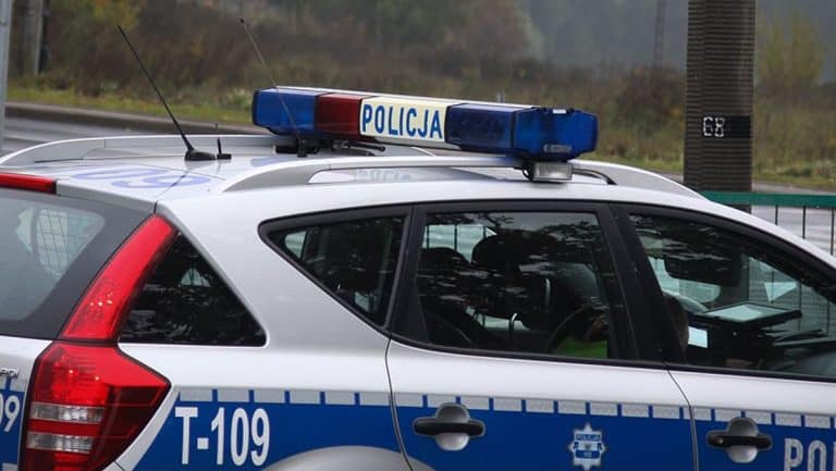 Policja na miejscu zdarzenia, samochód policyjny z włączonymi światłami, w tle drzewa i budynki.