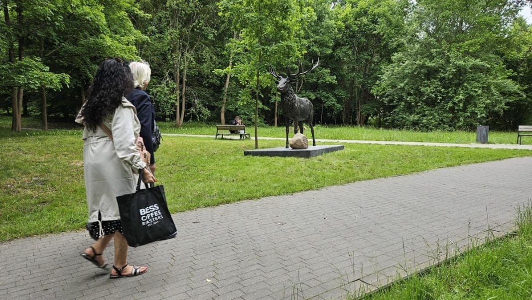Dwie kobiety patrzą na rzeźbę jelenia w parku, wokół dużo zieleni i ławki.
