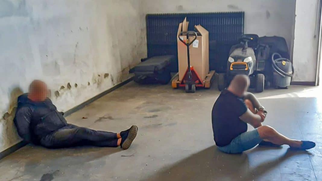 Dwoje osób siedzi na podłodze w pomieszczeniu, w tle narzędzia i sprzęt sprzątający.