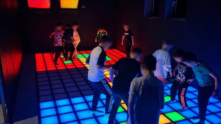 Grupa dzieci bawi się na interaktywnej macie LED w ciemnym pomieszczeniu, podświetlanej różnymi kolorami.