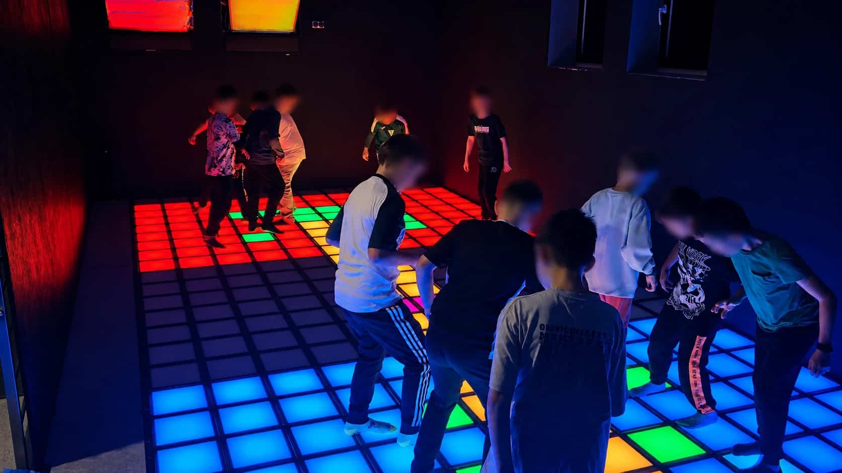Grupa dzieci bawi się na interaktywnej macie LED w ciemnym pomieszczeniu, podświetlanej różnymi kolorami.