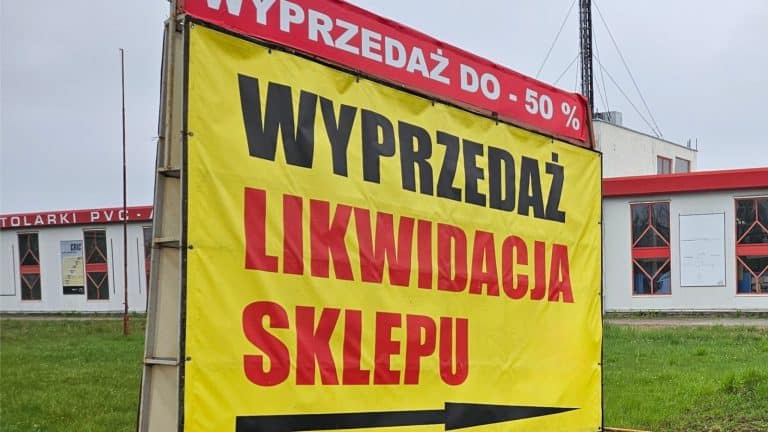 Baner informujący o likwidacji sklepu budowlanego z dużymi przecenami, na tle budynku z czerwonymi elementami.