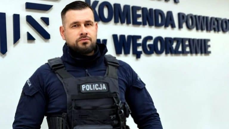 Policjant w mundurze z napisem 'Policja' na kamizelce, stojący przed budynkiem z napisem 'Komenda Powiatowa w Węgorzewie'.