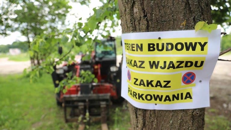 Tablica informująca o zakazie wjazdu i parkowania przy drzewie, w tle widoczny sprzęt budowlany.