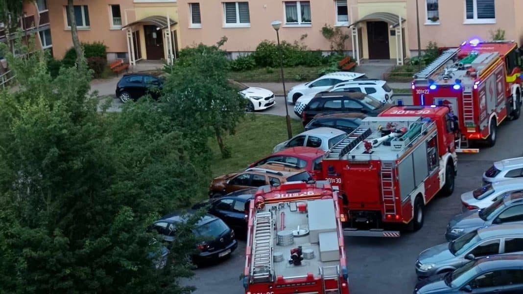 Dwa wozy strażackie i liczne samochody na osiedlowym parkingu, w tle bloki mieszkalne.
