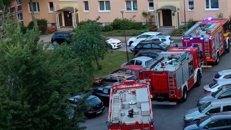 Dwa wozy strażackie i liczne samochody na osiedlowym parkingu, w tle bloki mieszkalne.
