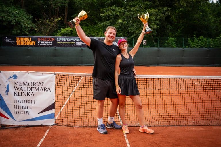 Dwóch uśmiechniętych tenisistów z trofeami na kortach w Olsztynie, stojący przy siatce.