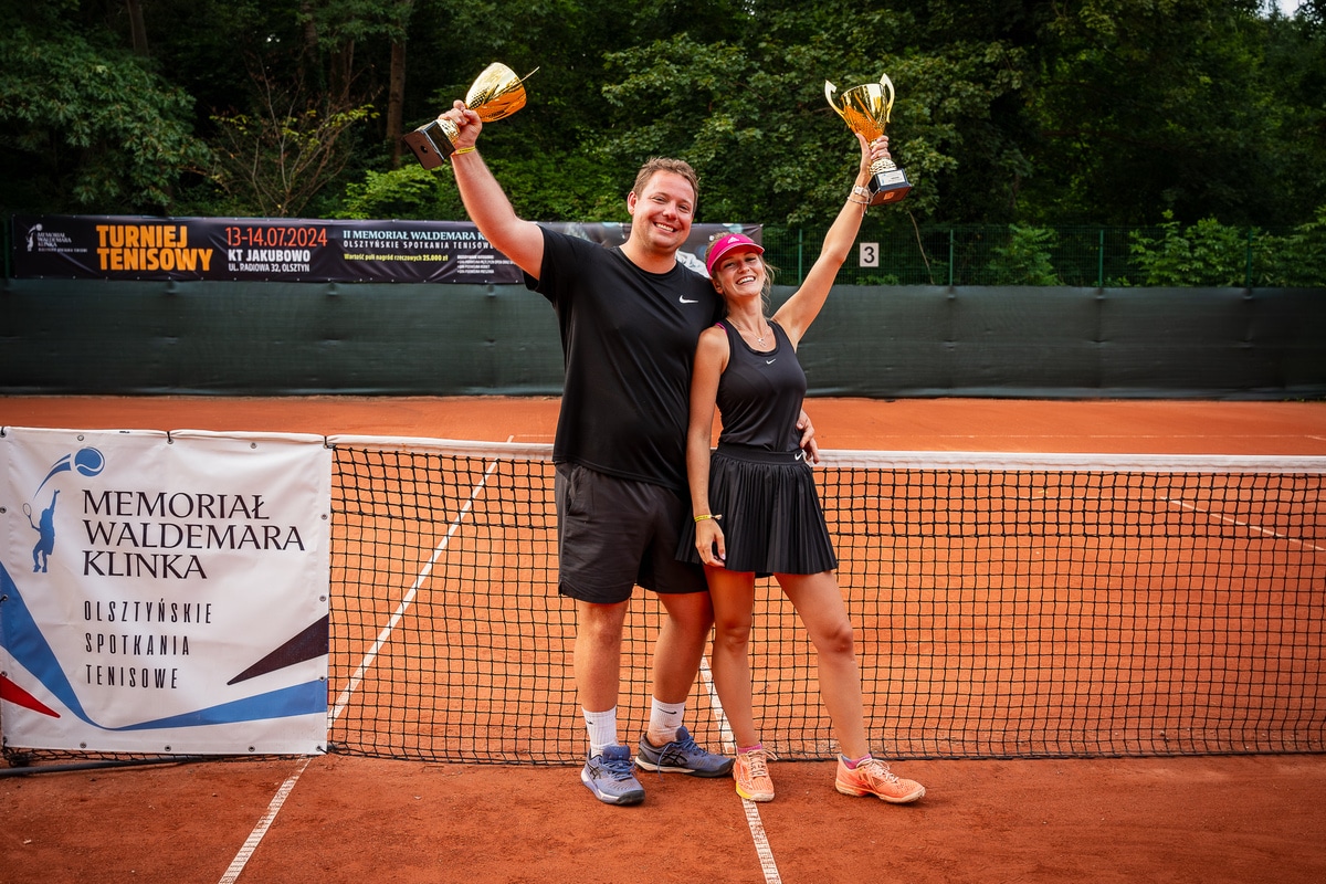 Dwóch uśmiechniętych tenisistów z trofeami na kortach w Olsztynie, stojący przy siatce.