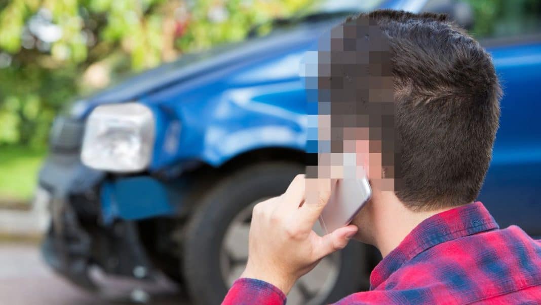 Mężczyzna rozmawia przez telefon obok uszkodzonego samochodu na tle zieleni.