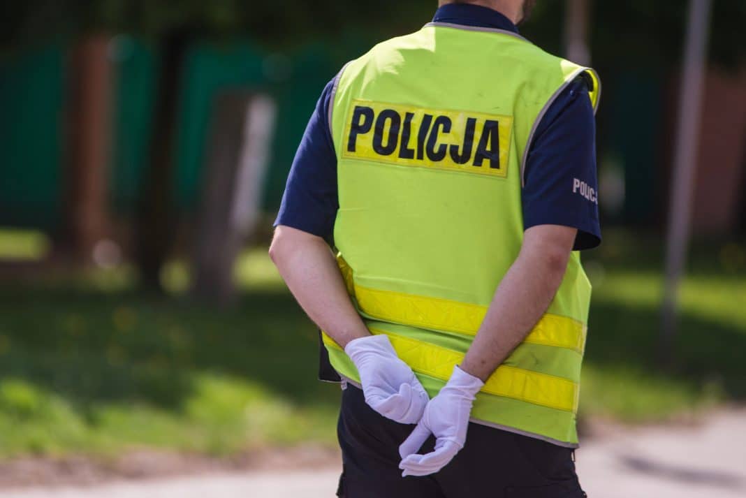 Policjant w kamizelce odblaskowej z napisem 'Policja' na tle zielonego otoczenia.